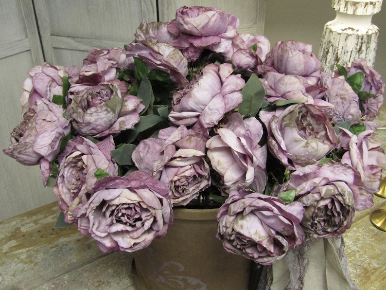 May include: A bouquet of dusty rose silk flowers arranged in a brown ceramic pot. The flowers are in full bloom and have a vintage aesthetic.