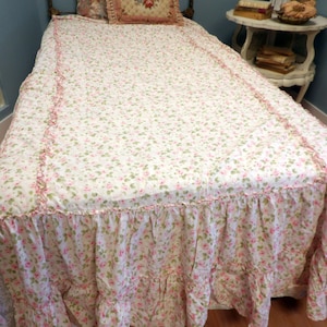 May include: A bed with a floral patterned bedspread and ruffled bed skirt. The bedspread is white with small pink and green flowers. A decorative pillow with a floral design rests on the bed. The bed frame is metal.