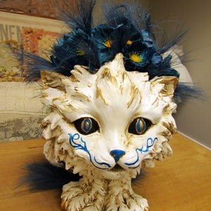 May include: White ceramic cat figurine with blue and gold painted details. The cat has a whimsical expression and is wearing a crown of blue flowers with yellow centers.