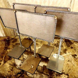 May include: A set of four rectangular silver-toned metal sign holders, each featuring a flat base and a vertical stand. The frames are decorated with a border of small, clear rhinestones and a single decorative rhinestone flower at the base.