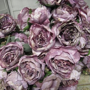 May include: A bouquet of artificial peonies in a dusty purple shade. The flowers are arranged in a cluster, with some petals overlapping. The peonies have a realistic appearance, with detailed textures and a slightly faded look.