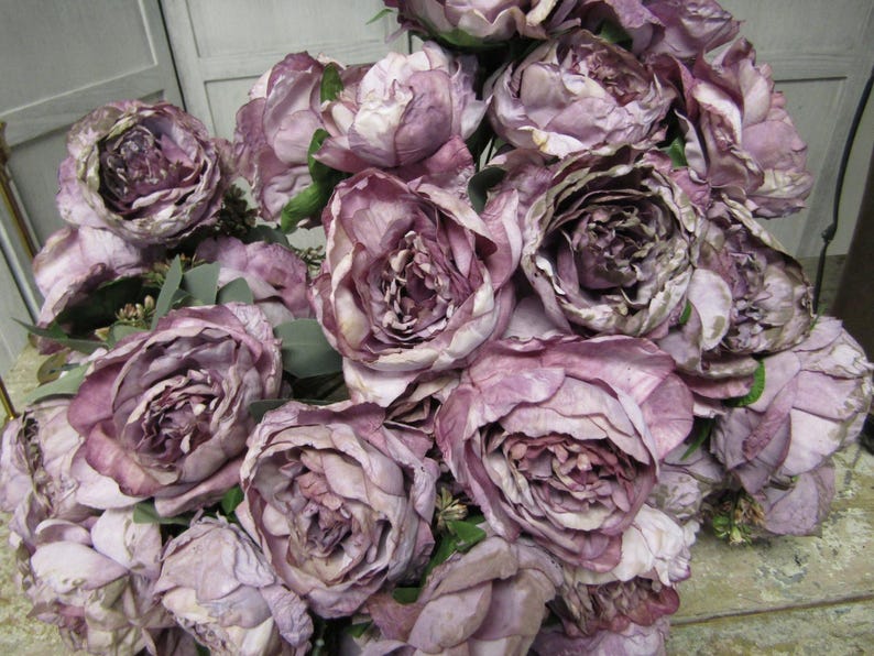 May include: A bouquet of artificial peonies in a dusty purple shade. The flowers are arranged in a cluster, with some petals overlapping. The peonies have a realistic appearance, with detailed textures and a slightly faded look.