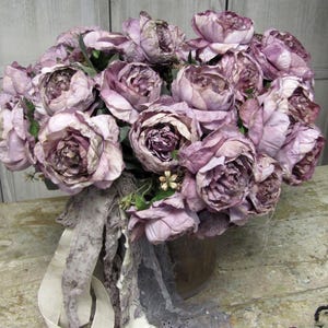 May include: A bouquet of dusty rose silk flowers arranged in a brown vase. The flowers are arranged in a loose, natural style, and the vase is decorated with a lace ribbon.