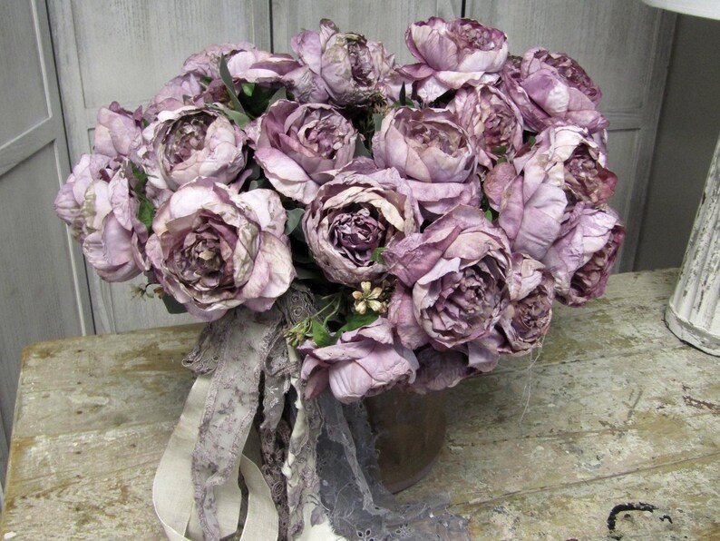 May include: A bouquet of dusty rose silk flowers arranged in a brown vase. The flowers are arranged in a loose, natural style, and the vase is decorated with a lace ribbon.