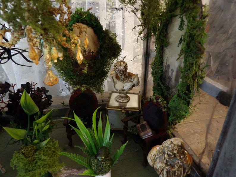 Op de afbeelding: Miniatuur diorama met een klassieke buste op een spiegeltafel, geflankeerd door kleine stoelen met donkerrode fluwelen zittingen. De sc&egrave;ne is versierd met groen, mos en hangende ornamenten, wat een grillige, antieke esthetiek cre&euml;ert.