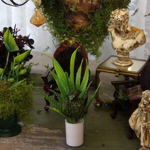 Op de afbeelding: Een decoratieve interieursc&egrave;ne met miniaturen. Een witte buste staat op een kleine glazen tafel. Kunstplanten in groene en donkerrode tinten zijn gerangschikt in kleine potten. Een kleine, donkere houten stoel is ook zichtbaar.