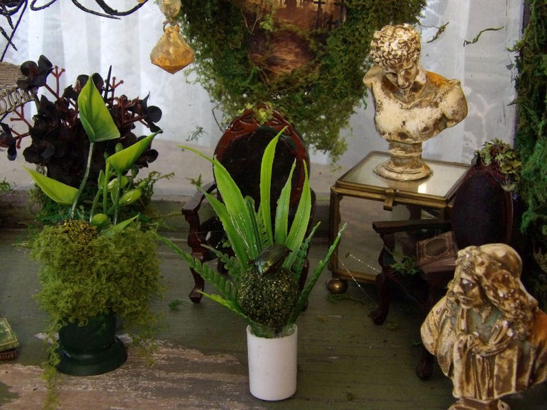 Op de afbeelding: Een decoratieve interieursc&egrave;ne met miniaturen. Een witte buste staat op een kleine glazen tafel. Kunstplanten in groene en donkerrode tinten zijn gerangschikt in kleine potten. Een kleine, donkere houten stoel is ook zichtbaar.