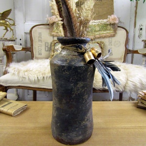 May include: A dark brown ceramic vase with a rustic finish, filled with dried feathers and bells. The vase is sitting on a wooden table.
