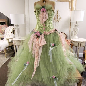 May include: A green tulle ball gown with pink and white lace accents and a green and gold brocade bodice. The gown is decorated with pink roses and has a crown on top.