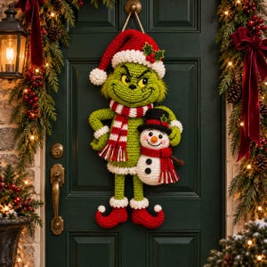 Könnte beinhalten: Ein gehäkelter Wandbehang, der eine grüne Figur mit schelmischem Gesichtsausdruck zeigt, die eine rot-weiße Weihnachtsmütze und einen passenden gestreiften Schal trägt. Die Figur hält einen kleinen weißen Schneemann mit schwarzem Zylinder.