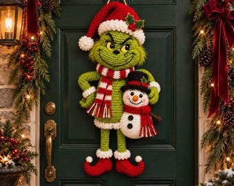 May include: A crocheted wall hanging featuring a green character with a mischievous expression, wearing a red and white Santa hat and a matching striped scarf. The character holds a small white snowman with a black top hat and a red scarf.