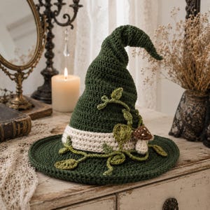May include: A dark green crocheted pointed hat featuring a wide brim and a cream-coloured band around the base. The hat is decorated with crocheted green vines, leaves, and a small brown and white mushroom attached to the side of the crown.