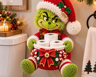 May include: A crocheted Grinch-inspired toilet paper holder featuring a green character wearing a red Santa hat with a white pom-pom. The figure holds a red basket decorated with a large red bow, gold bell, and candy canes, containing three rolls of toilet paper.