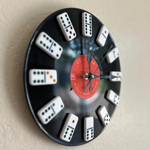May include: A circular wall clock crafted from a black vinyl record featuring white dominoes as hour markers. The center label is red with white text reading Columbia and Mono. The clock has three black hands positioned at the center of the record.