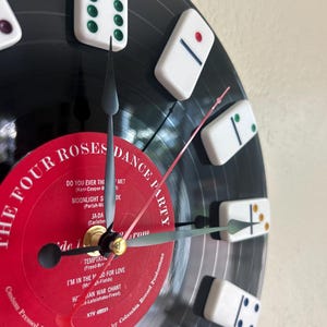 May include: A wall clock made from a black vinyl record with a red center label reading THE FOUR ROSES DANCE PARTY. White dominoes with colored pips serve as hour markers. The clock features black hour and minute hands and a thin red second hand.