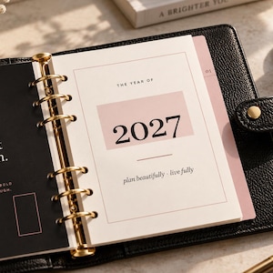 May include: A black leather binder with gold rings holding white pages. The left page is black with the text "Make it happen." The right page is white with a pink rectangle, the text "THE YEAR OF 2027", and the phrase "plan beautifully · live fully".