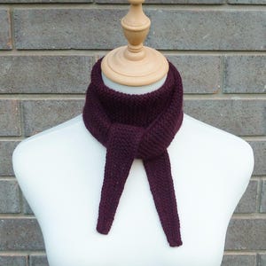 May include: A knitted scarf in a deep burgundy color is displayed on a mannequin. The scarf is made of a thick yarn, with a textured appearance. The scarf is tied in a knot, with the ends hanging down. The background is a brick wall.