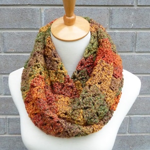 May include: A chunky crochet infinity scarf featuring a variegated yarn pattern in autumn-inspired shades of burnt orange, deep forest green, golden yellow, and dark brown. The textured knit design consists of repeating openwork clusters.
