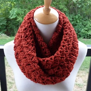 May include: A rust-colored, crocheted infinity scarf displayed on a mannequin. The scarf has a textured, open weave design. The warm, earthy tone of the scarf suggests it is suitable for fall or winter wear. The scarf is a circular loop, designed to be worn around the neck.