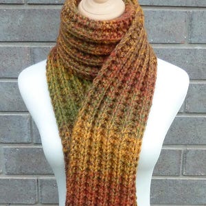 May include: A long, thick, hand-knitted scarf featuring a ribbed texture and a variegated colour palette of autumn tones, including deep forest green, burnt orange, mustard yellow, and dark brown, wrapped around a white mannequin neck.
