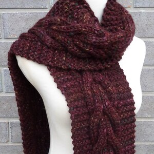 May include: A maroon knitted scarf with a cable knit pattern. The scarf is displayed on a white mannequin.