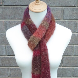 May include: A long, fuzzy, knitted scarf featuring a variegated color palette of deep burgundy, muted brown, and hints of purple. The scarf is wrapped around a mannequin neck, displaying a soft, textured yarn construction with a loose, open knit.