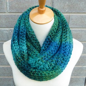 May include: A teal, green and blue infinity scarf made of yarn. The scarf is displayed on a white mannequin.