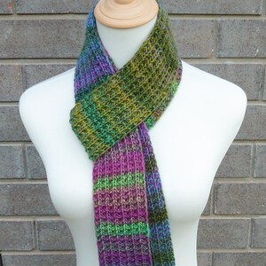 May include: A colorful knitted scarf displayed on a mannequin. The scarf features a ribbed pattern and a gradient of colors, including purple, green, and blue. The scarf is made of soft yarn and is suitable for cold weather.