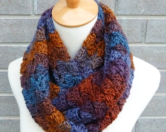May include: A crocheted infinity scarf in shades of brown, blue, and purple. The scarf is made with a textured stitch and is displayed on a white mannequin.