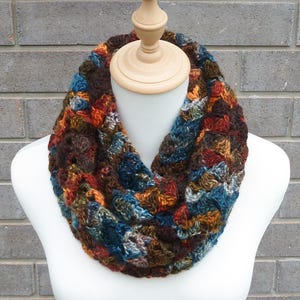 May include: A handmade, multicoloured crochet infinity scarf. The scarf features a mix of brown, blue, orange, and red yarn, creating a textured, warm accessory. The scarf is displayed on a white mannequin, showcasing its circular design.