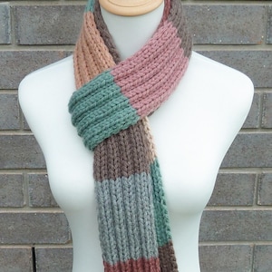 May include: A knitted scarf in a variety of colours, displayed on a mannequin. The scarf has a ribbed texture and is divided into sections of pink, green, brown, and grey. The scarf is made of a soft, warm material, perfect for colder weather.