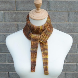 May include: A hand-knitted neck scarf featuring a ribbed texture and a variegated color palette of earthy tones including brown, mustard yellow, olive green, and deep orange. The scarf is wrapped around the neck with two ends hanging down.