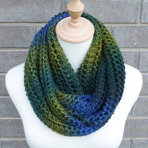 May include: A knitted infinity scarf with a gradient of blue, green and black colours. The scarf is made with a textured stitch and is displayed on a white mannequin.