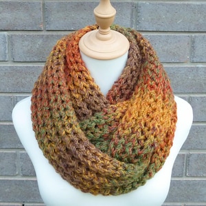May include: A chunky knit infinity scarf featuring a variegated yarn pattern with blended tones of burnt orange, deep brown, forest green, and golden yellow. The scarf has a thick, open-stitch texture and is displayed on a neutral mannequin form.