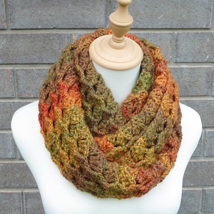May include: A chunky crochet infinity scarf featuring a variegated yarn pattern in earthy tones of forest green, burnt orange, golden yellow, and deep brown. The textured, looped design creates a thick, layered appearance around the neck.