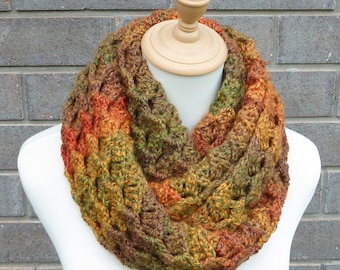 May include: A chunky crochet infinity scarf featuring a variegated yarn pattern in earthy tones of forest green, burnt orange, golden yellow, and deep brown. The textured, looped design creates a thick, layered appearance around the neck.