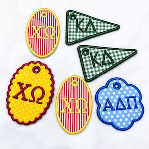 May include: A collection of six embroidered fabric patches featuring Greek letters. The set includes oval, triangular, and scalloped shapes in red and white stripes, green and white gingham, yellow with white dots, and blue with white dots.
