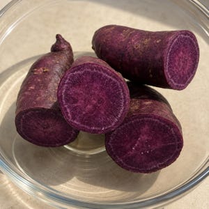 May include: Several pieces of raw purple sweet potato are arranged inside a clear glass bowl. The tubers have a dark purple skin and a vibrant, deep purple interior flesh that is visible where the potatoes have been sliced into thick rounds.