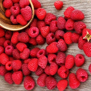 May include: A collection of fresh, vibrant red raspberries scattered across a textured wooden surface, with a portion of the fruit contained within a small, round, light-brown wooden bowl positioned in the upper left corner of the frame.