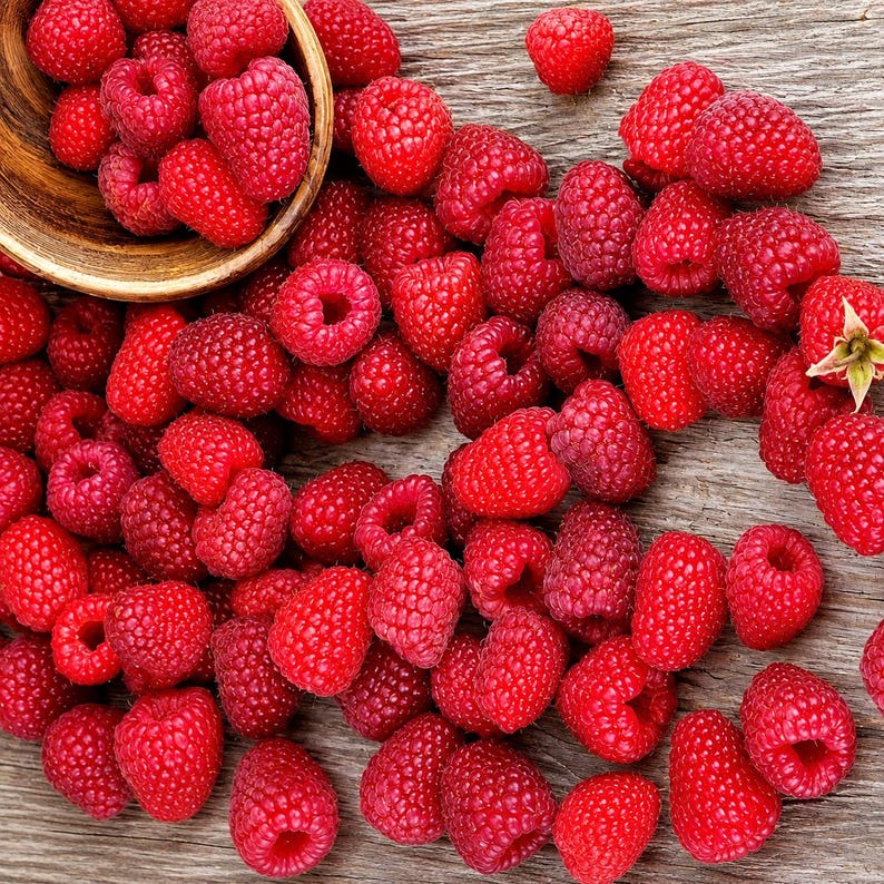 May include: A collection of fresh, vibrant red raspberries scattered across a textured wooden surface, with a portion of the fruit contained within a small, round, light-brown wooden bowl positioned in the upper left corner of the frame.