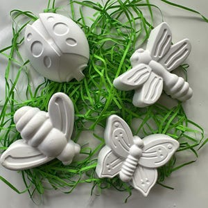 May include: Four white, moulded insect-shaped figures including a ladybird, a bee, and two butterflies. Each figure features raised, smooth surfaces and distinct anatomical details, arranged together on a bed of thin, bright green paper strips.
