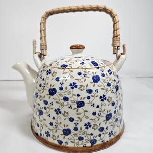 May include: A ceramic teapot with a cream-coloured base featuring a repeating pattern of small blue roses and brown vines. The teapot has a curved spout, a matching lid with a brown knob, and a swing handle wrapped in light brown woven rattan.