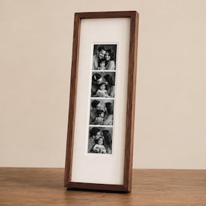 May include: A tall, rectangular dark wood picture frame featuring a white mat board with four vertical openings. Each opening contains a black and white photograph of a family consisting of two adults and one child in various poses.