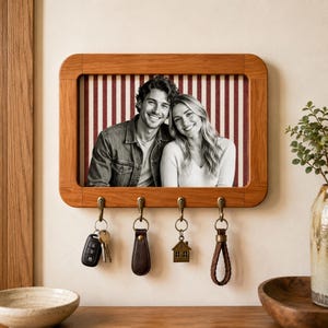 May include: A rectangular wooden wall-mounted key holder featuring a central photo frame displaying a black and white portrait of a smiling couple against a red and white striped background. Four brass-toned metal hooks hang from the bottom edge.