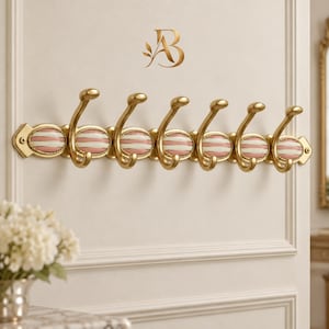 May include: A wall-mounted coat rack featuring six gold-toned metal hooks. Each hook is attached to an oval-shaped base with horizontal pink and white stripes. The rack is secured with screws at both ends and displays a gold logo above it.