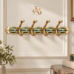 May include: A wall-mounted coat rack featuring a gold-toned metal base with four curved hooks. The rack includes five oval-shaped ceramic inserts decorated with horizontal dark green and white stripes, secured by screws at both ends.