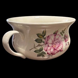 May include: A cream-colored ceramic chamber pot featuring a single side handle and a wide, flared rim. The exterior is decorated with a detailed botanical illustration of a large pink rose blossom surrounded by green leaves and smaller rosebuds.