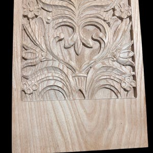 May include: A rectangular light brown wooden panel featuring an intricate hand-carved relief design. The carving displays symmetrical floral and leafy scrollwork patterns centered around a heart-shaped motif, with a smooth, uncarved lower section.