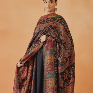 May include: A black shawl featuring intricate, multi-colored woven patterns in shades of pink, teal, gold, and orange. The design includes detailed figurative motifs of people and animals throughout the fabric, finished with a decorative border.