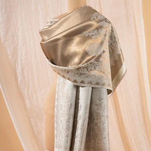Puede incluir: Un chal rectangular con una paleta de colores dorado metálico y crema, con intrincados patrones de bordado floral y de cachemira en blanco. La tela tiene un acabado suave y brillante y está drapeada sobre un maniquí para mostrar el borde.